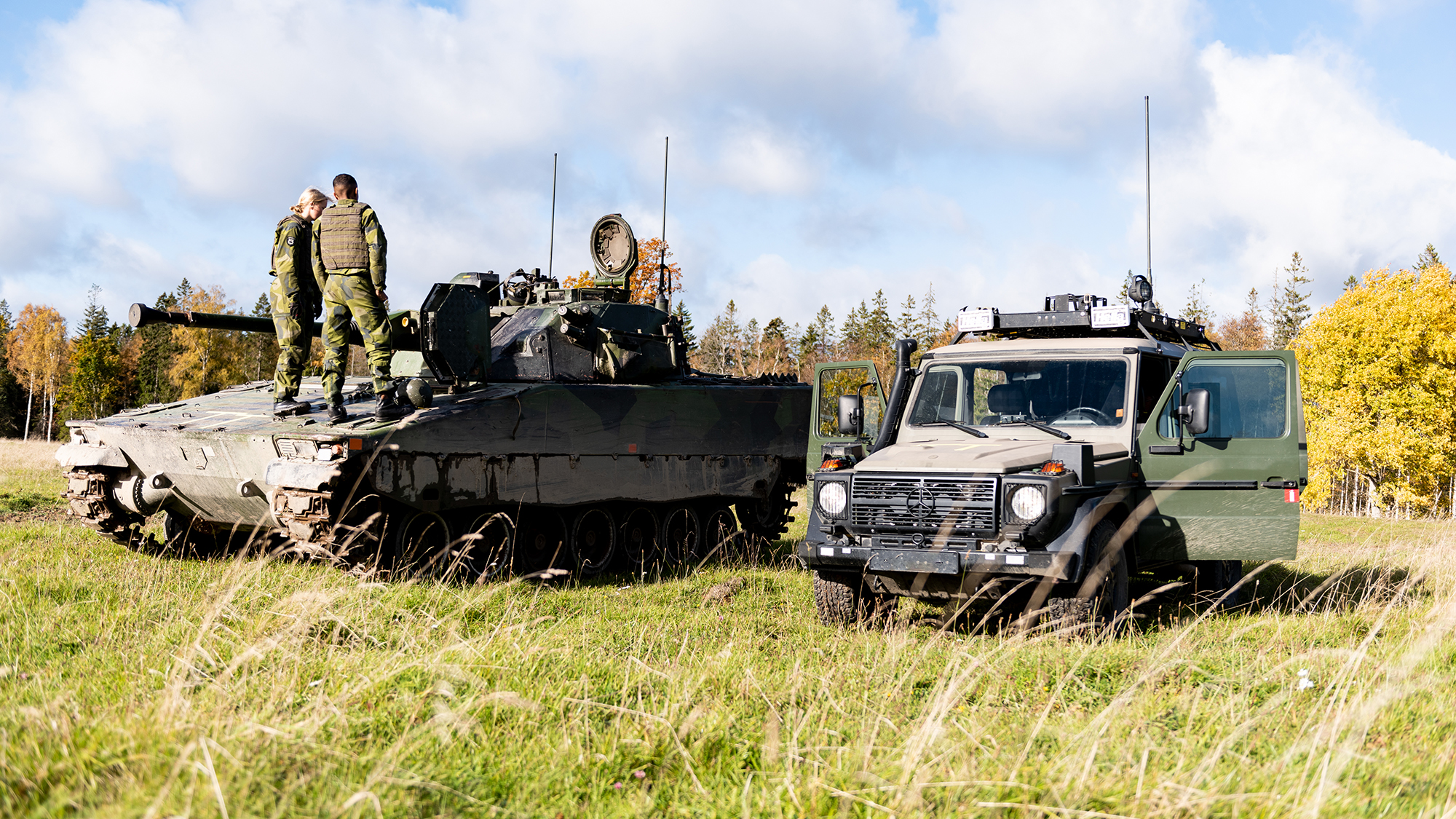 Två personer står på ett stridsfordon. Ett fordon står parkerat bredvid.