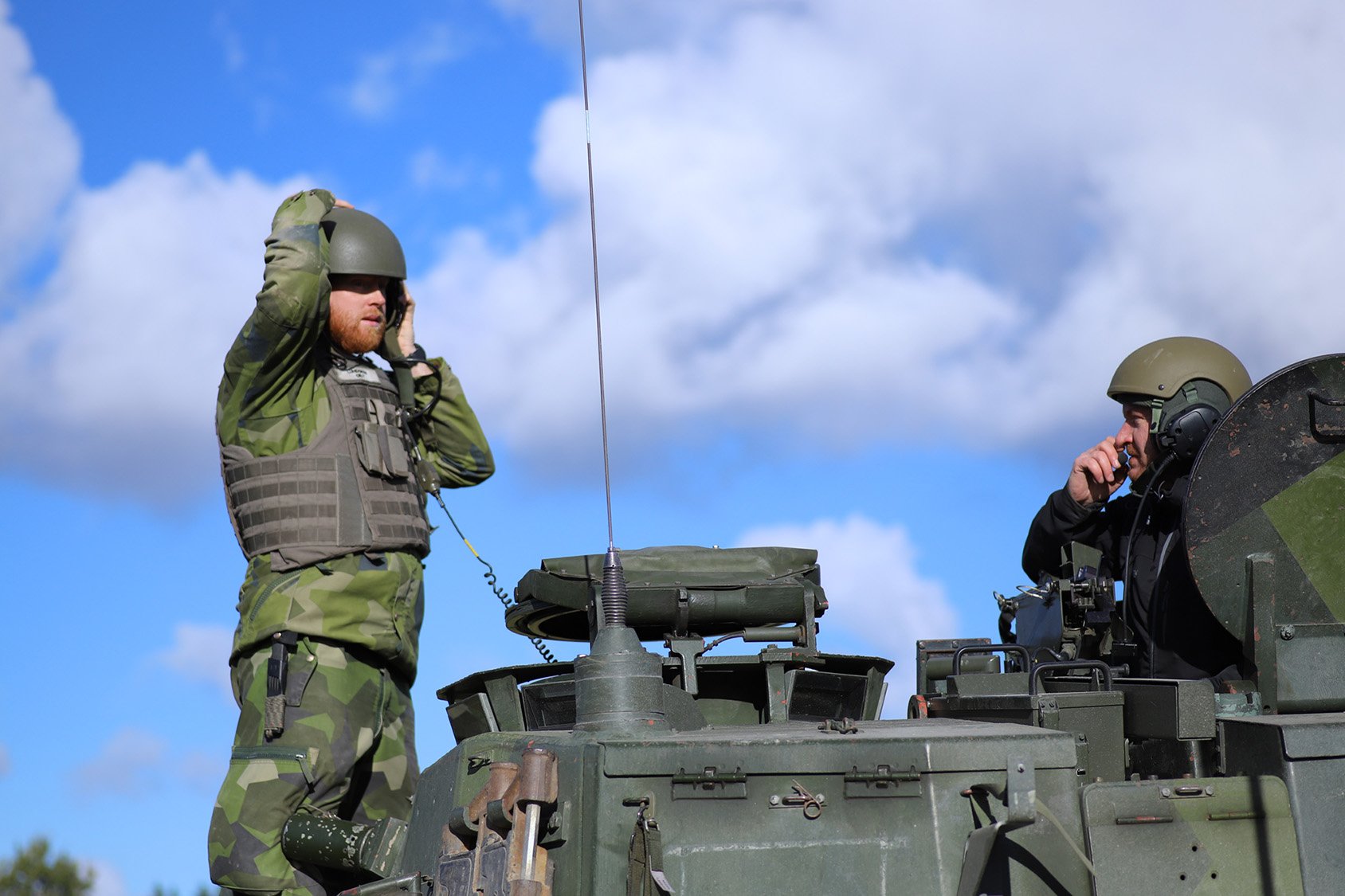 Två soldater står på ett stridsfordon och pratar i kommunikationsradio.